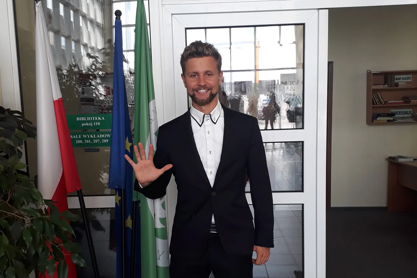 Adam Gajda w garniturze na uczelni, macha do aparatu